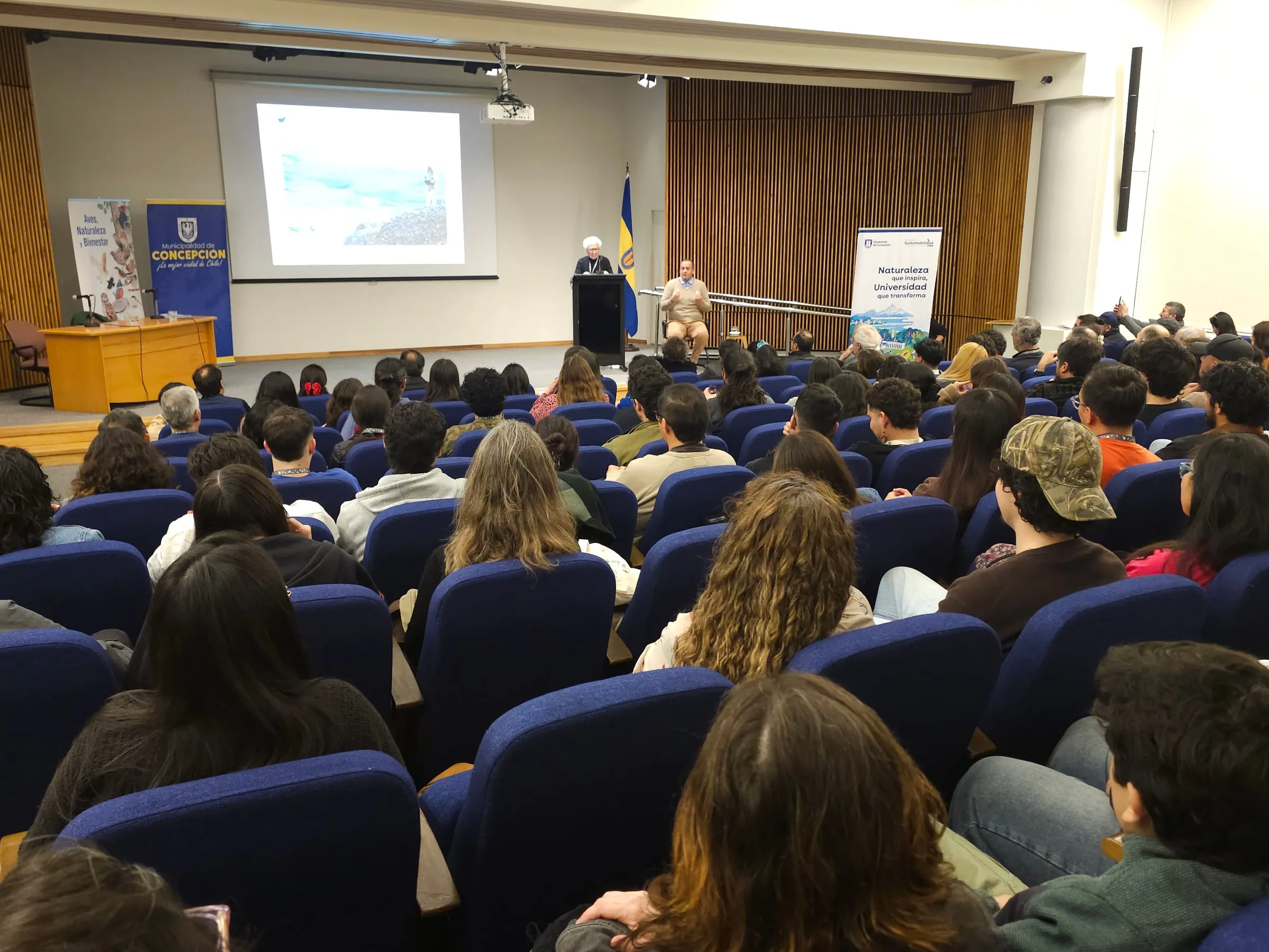 Auditorio lleno durante la conferencia inaugural, con una persona en el podio y un intérprete de lengua de señas.