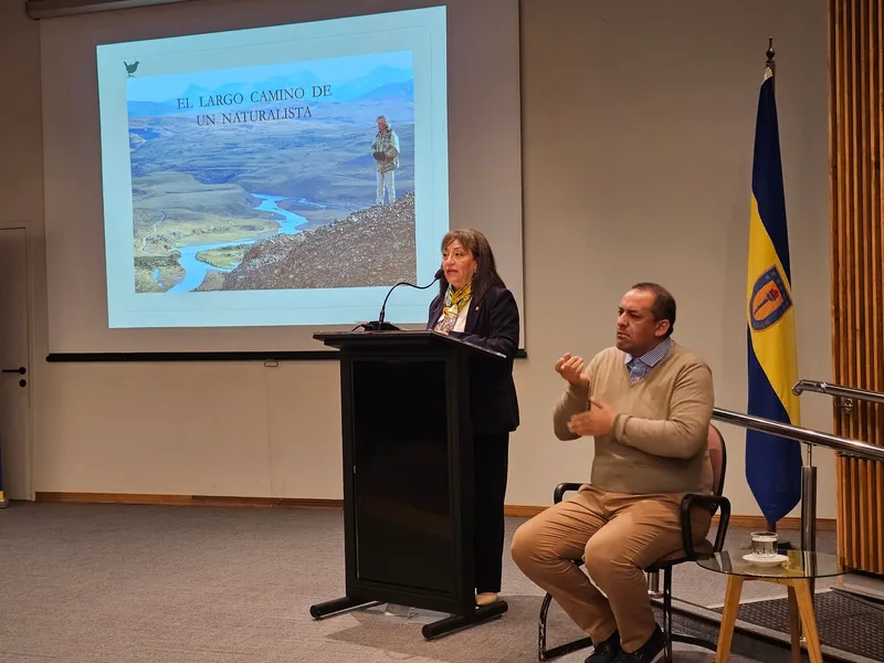 Una mujer habla desde un podio frente a la proyección «El largo camino de un naturalista», junto a un intérprete de lengua de señas.