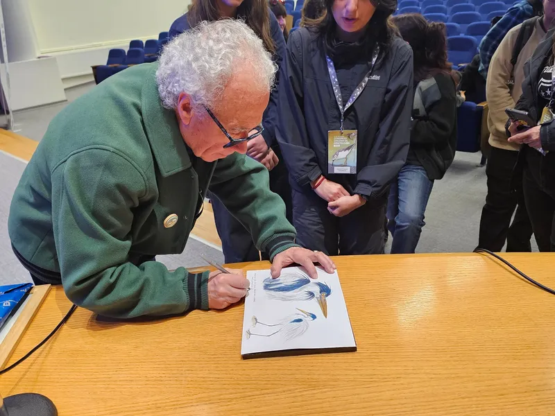 Un hombre de cabello blanco firma un libro con ilustraciones de garzas, rodeado de estudiantes.