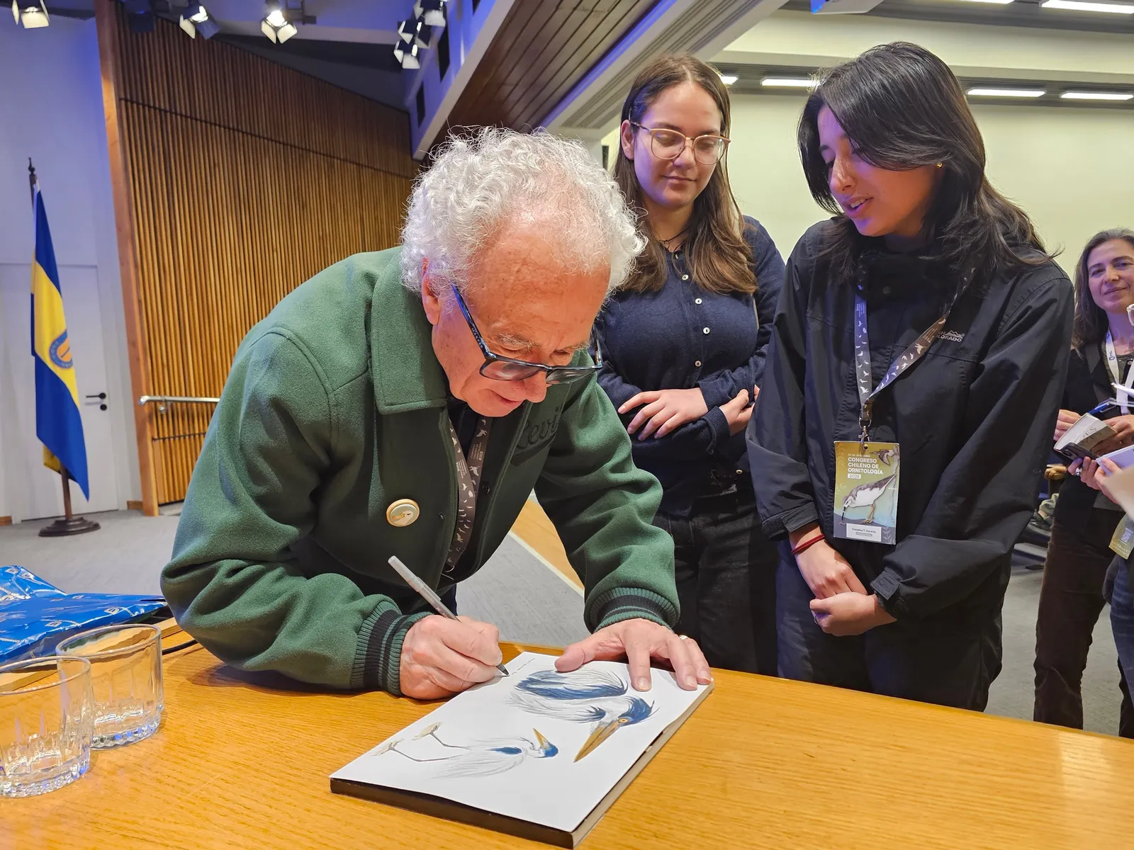 Un hombre de cabello blanco firma un libro de ilustraciones de aves mientras dos jóvenes observan.