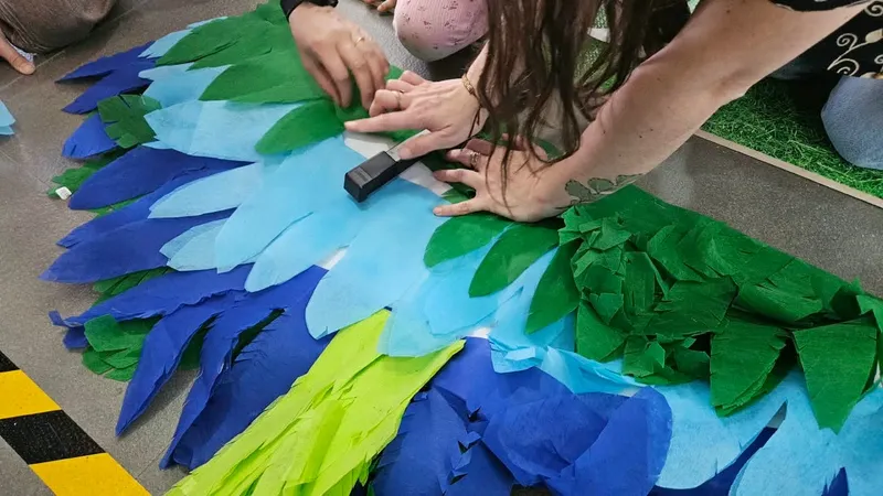 Manos que arman unas alas con plumas de papel azul, celeste y verde, sobre el suelo.